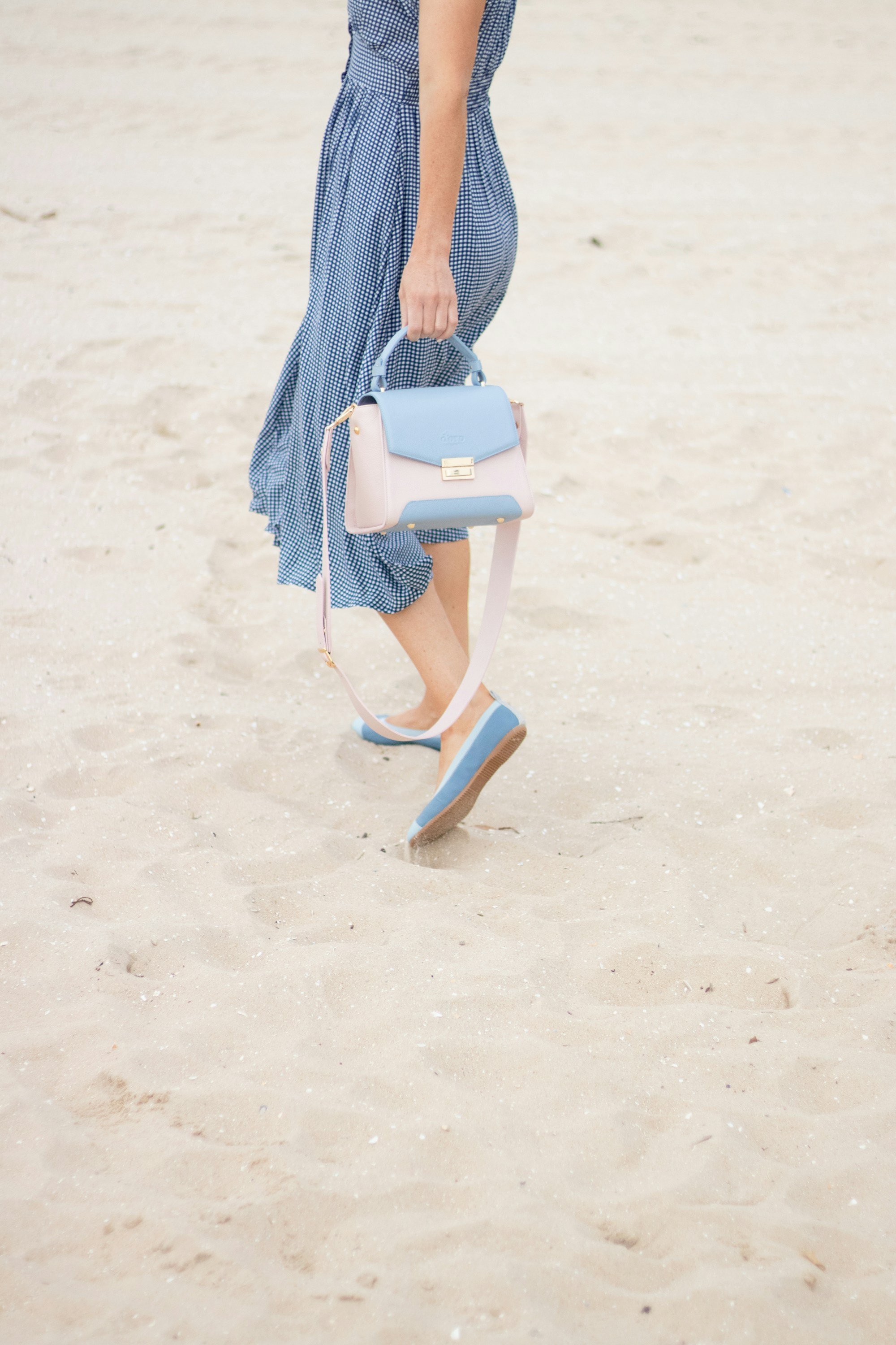 A woman in a blue polka-dot midi dress walks across the sand carrying a small structured leather top-handle bag