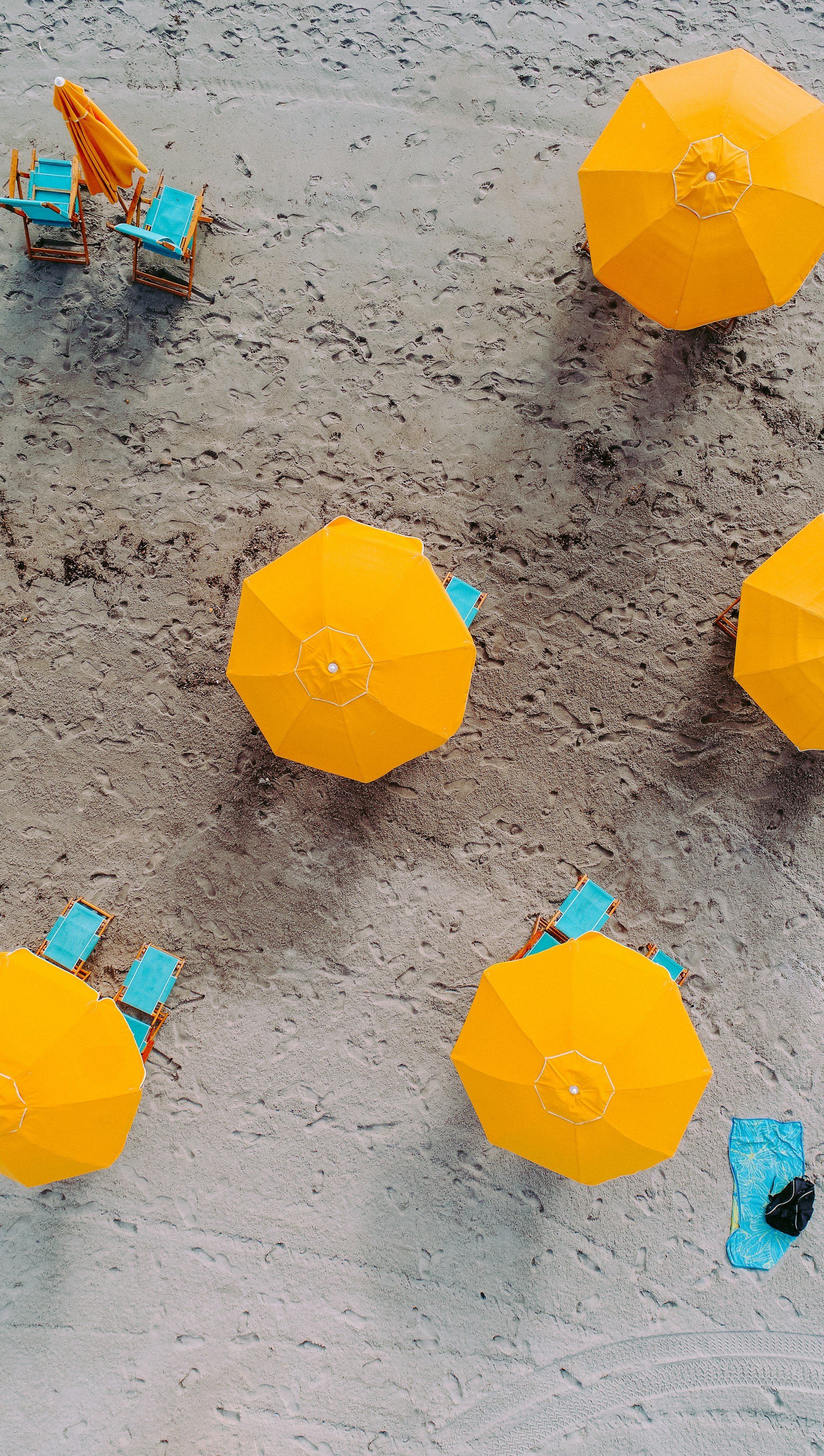 Aerial view of yellow beach umbrellas with pairs of teal chairs casting long shadows across the sand, one umbrella closed
